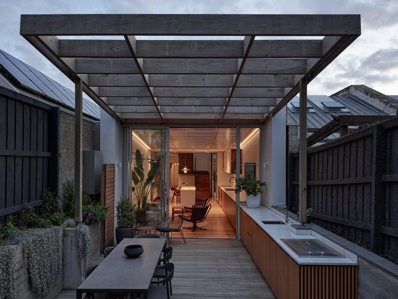 Rear courtyard with pergola, outdoor sink, and decking leading to an illuminated interior at dusk