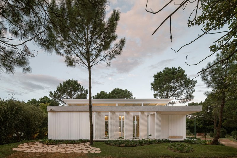Street view of the white ribbed facade with flat cantilevered roof framed by tall pines