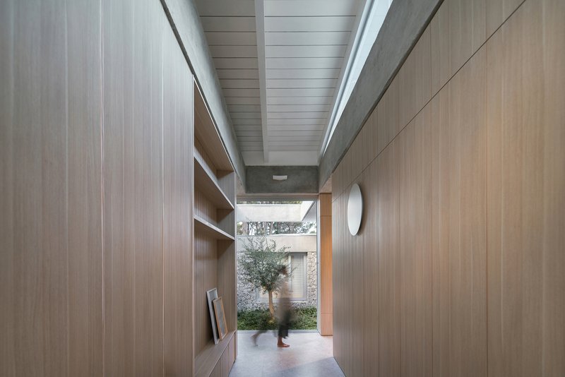Hallway with timber-lined walls and ceiling leading to a planted courtyard as a figure passes through