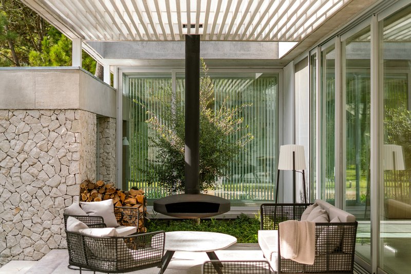 Covered terrace with slatted pergola, suspended fireplace, and stone wall beside planted olive tree