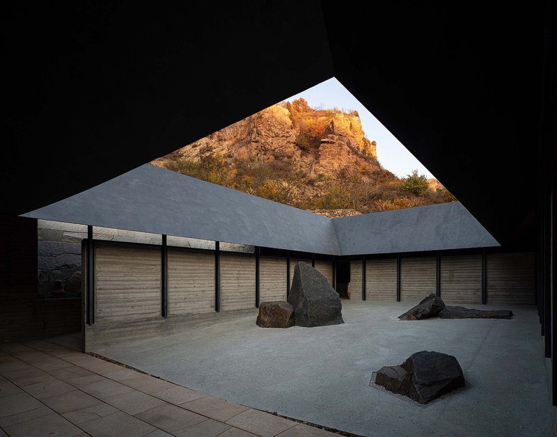 Interior courtyard framed by angular roof opening revealing weathered rock face illuminated by warm afternoon light