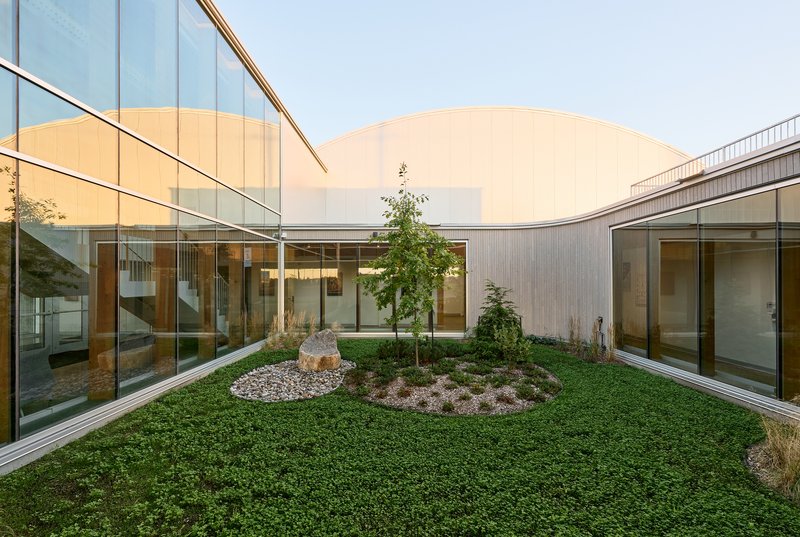 Planted courtyard with circular groundcover bed and trees surrounded by glass-walled corridors under an overcast sky