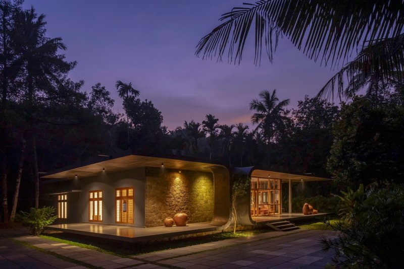 Curved roofline with illuminated stone wall and glazed entrance at dusk beneath palm trees