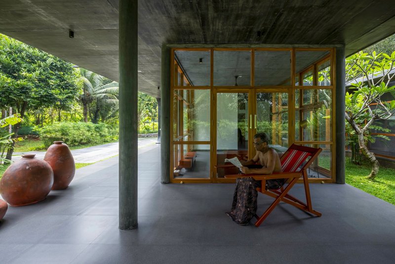 Covered terrace with concrete columns and timber-framed glazing opening to tropical garden with person reading