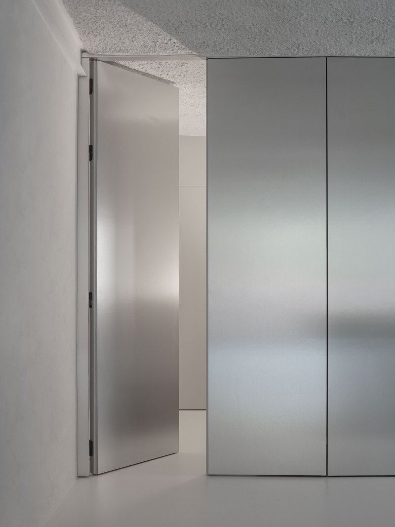 Brushed steel pivot door opening against floor-to-ceiling cabinet panels in soft grey light