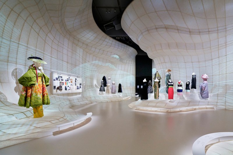Curved white grid-patterned walls enclosing a gallery space with fashion mannequins on display platforms
