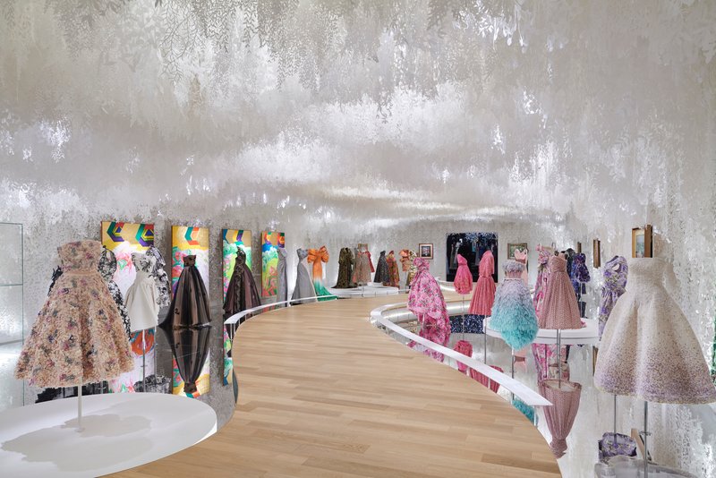 Curved exhibition gallery with textured silver-leaf wall finish and light wood flooring displaying couture garments