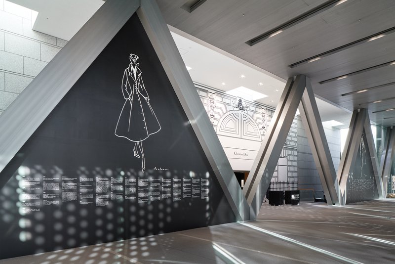 Exhibition corridor with diagonal concrete columns framing a black display wall and ornate facade beyond