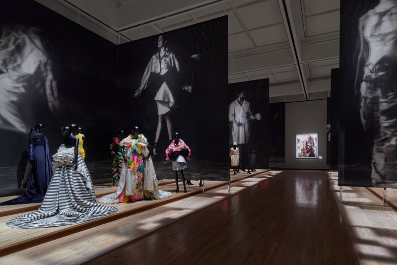 Exhibition room with dark walls showing projected images above mannequins and garments on wooden floor