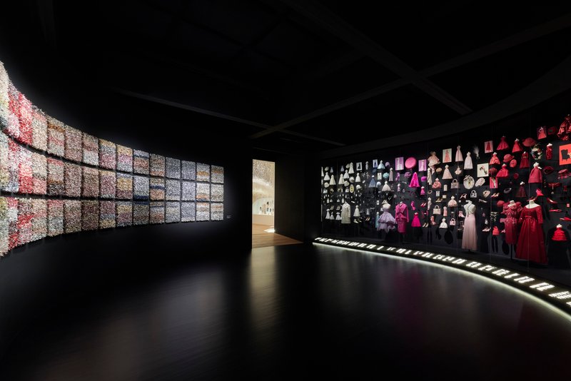 Curving exhibition space with backlit image grid and wall of garments under a black coffered ceiling
