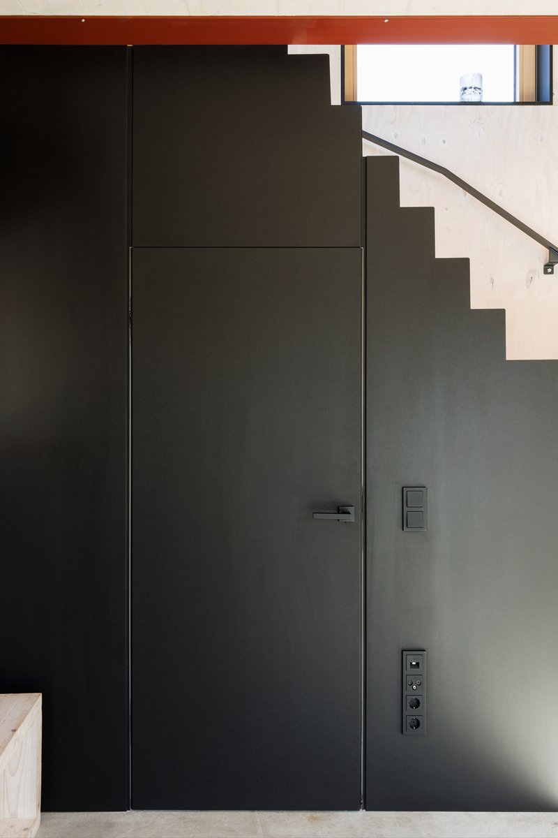 Flush black metal door set into grey wall beneath visible stair treads and red ceiling beam