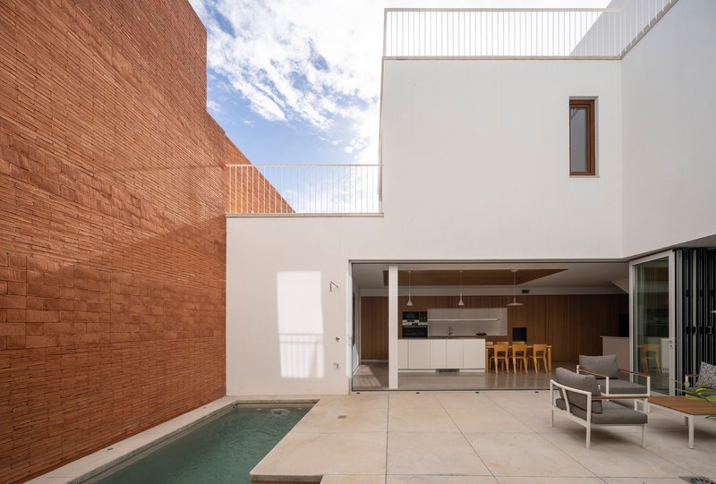 Courtyard with narrow pool flanked by brick wall and white rendered volumes under blue sky