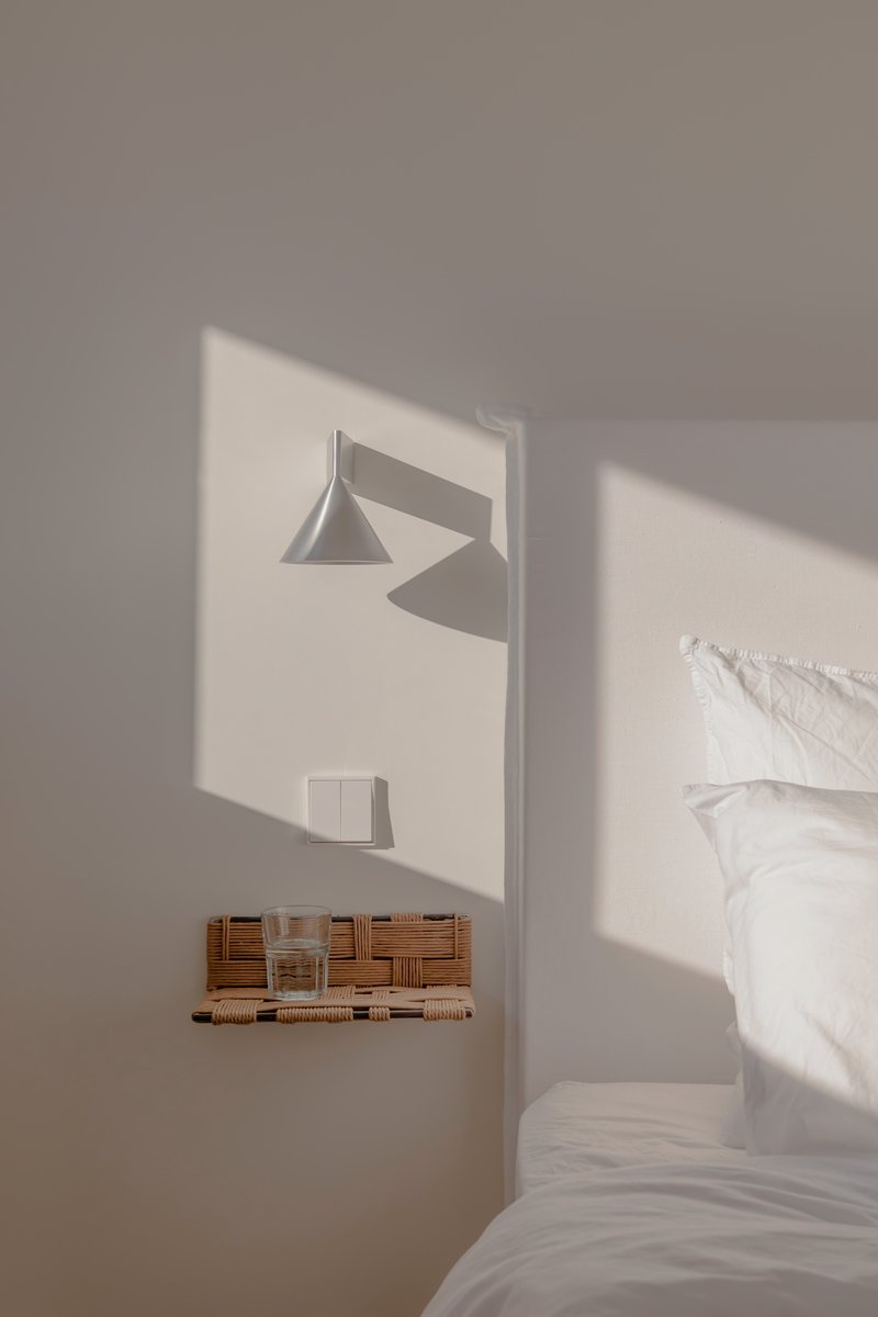 Bedroom niche with wall-mounted lamp and woven shelf casting afternoon sunlight across white surfaces