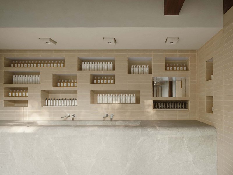 This architectural image showcases a modern bathroom interior with a minimalist design. The space features a beige tile wall with recessed shelves displaying va