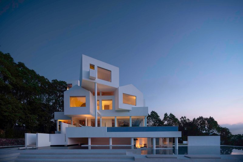 White stacked volumes with illuminated rectangular window openings at dusk beside a reflecting pool
