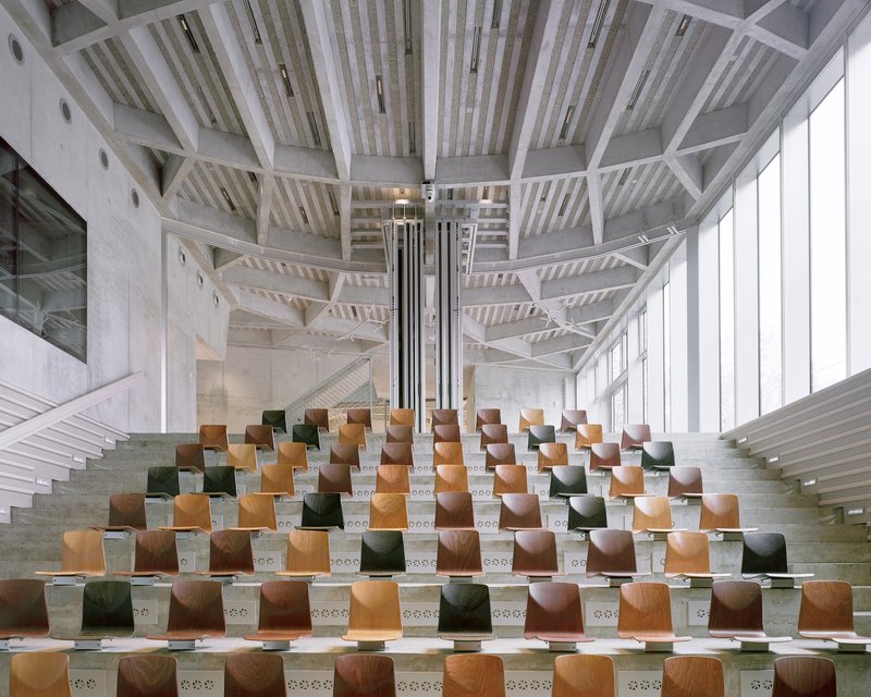 Interior tiered seating with colored wooden seats arranged beneath exposed concrete beams and skylights