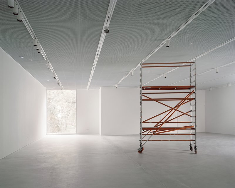 White gallery space with polished concrete floor, track lighting, and a rolling scaffold near the window