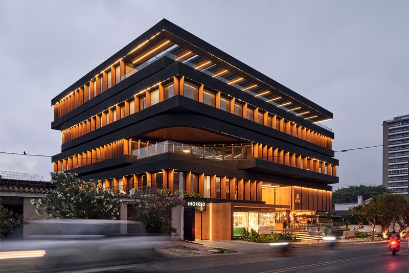 Corner view showing the terraced facade with illuminated timber fins and ground-level retail at twilight