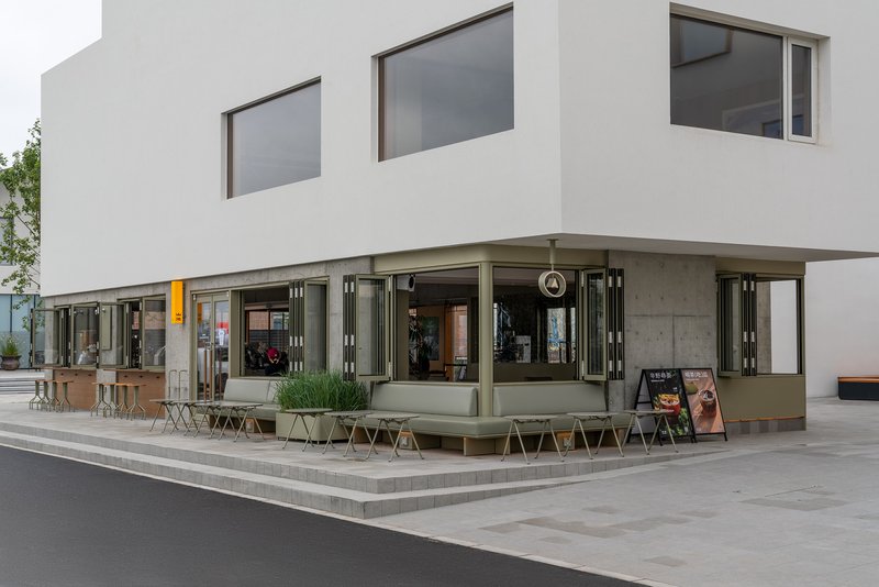 Corner detail of the folding glass storefront with olive-green frames and outdoor seating on the plaza