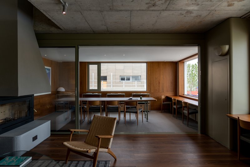 Interior view through wood-paneled spaces with exposed concrete ceiling and woven chair in foreground