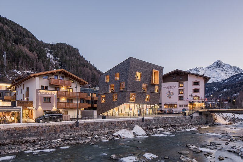 Cubic volume clad in dark shingles nestled between traditional alpine buildings along a winter stream