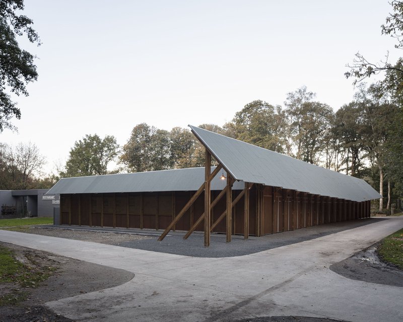 Long pavilion with timber portal frames and metal roofing beside a paved path on an overcast autumn day