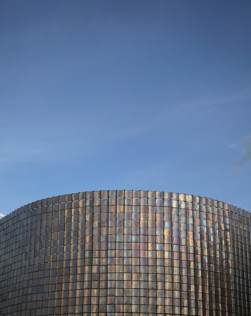 Curved facade clad in iridescent glass tiles catching late afternoon light against a clear sky