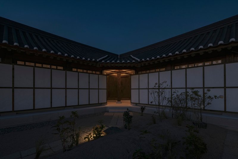 Interior courtyard with translucent wall panels and planted beds beneath converging tiled rooflines at twilight