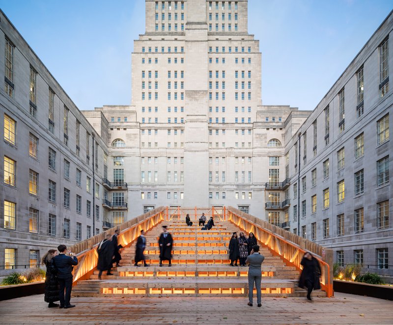 Illuminated timber and glass staircase bridging a courtyard between stone facades at dusk