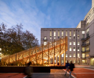 The image showcases a contemporary wooden pavilion in an urban setting, juxtaposed against a classic