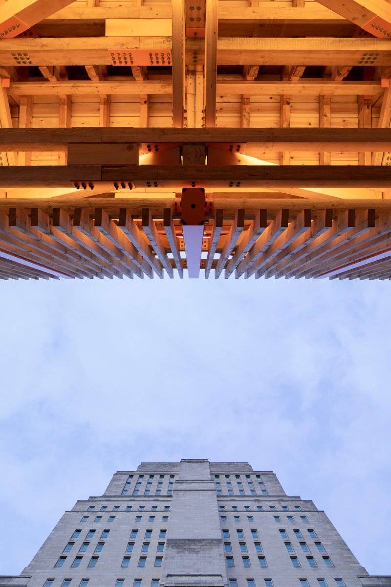 Upward view through layered timber joists and beams with a stepped concrete tower beyond