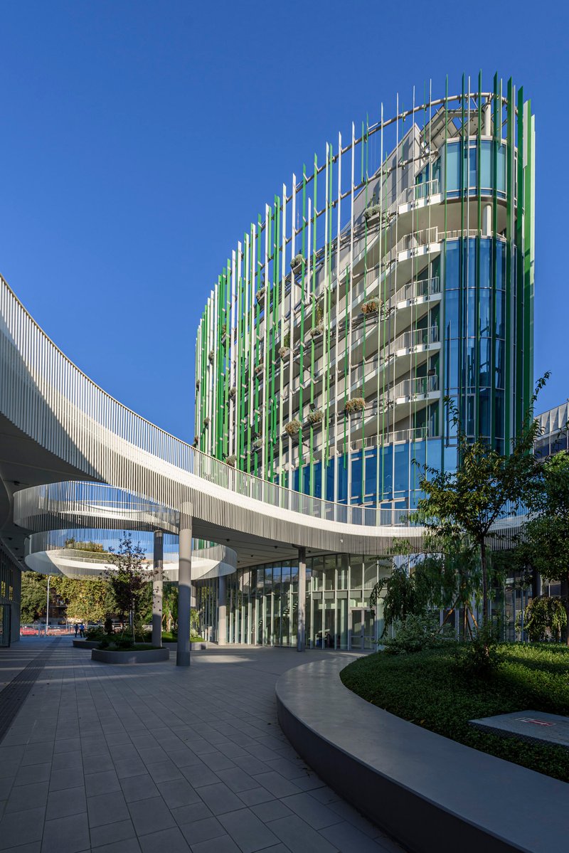 Ground-level view of the glazed tower with vertical fin screen and elevated walkway spanning above planted beds