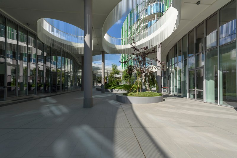 Covered courtyard beneath circular roof openings with central planted island and glazed perimeter walls casting shadows