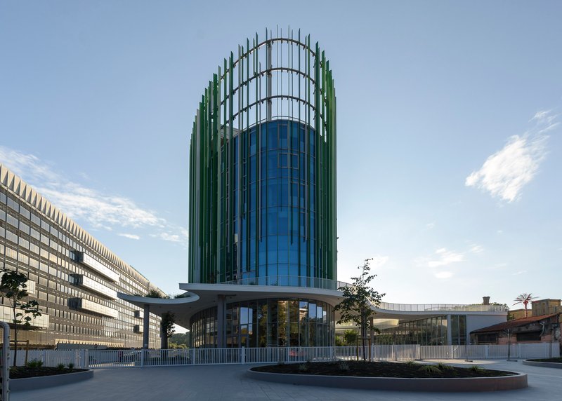 Cylindrical glass tower with vertical fin cladding viewed from the plaza with elevated pedestrian bridge at base