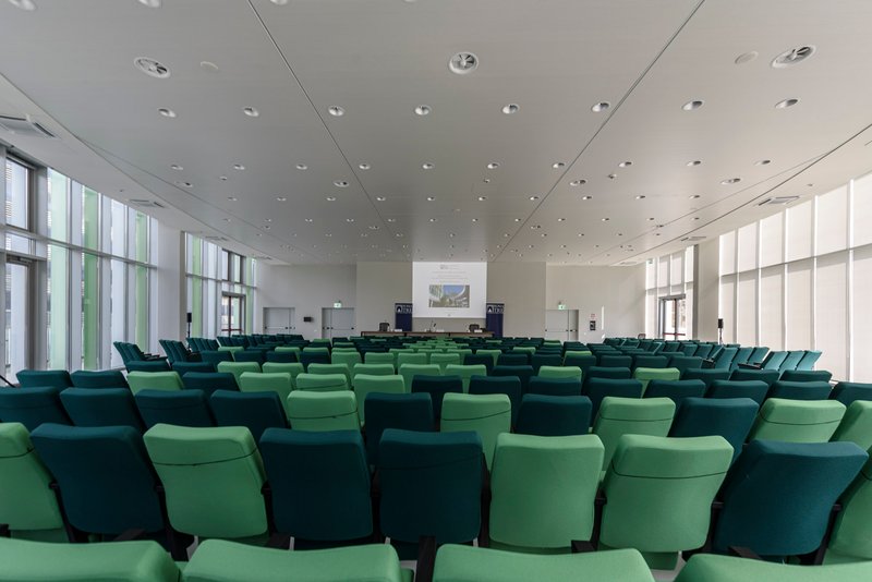 Auditorium interior with rows of green upholstered seating beneath white ceiling panels and recessed lighting