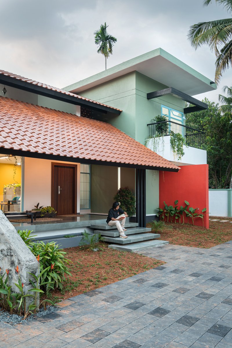 Street facade showing terracotta roof, coral red wall, and resident on entrance steps under palms
