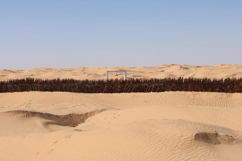 Distant view of a freestanding portal frame beyond a palm frond fence in rolling sand dunes