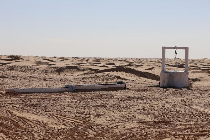 Concrete well structure with pulley system and adjacent water trough surrounded by tire tracks in sand