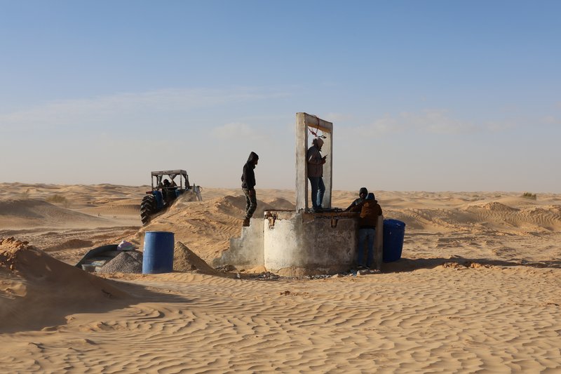 Concrete well structure with vertical frame opening and blue barrels amid windswept desert sand