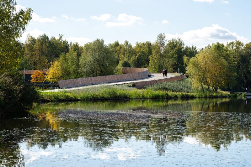 View across the water of the elevated timber walkway threading through planted wetland grasses with two figures strolling