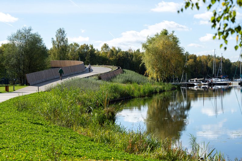 The elevated promenade curving past waterside planting beds with sailboats moored beyond on a sunny afternoon