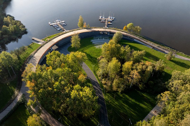 Aerial view of the curved pedestrian bridge enclosing a wooded island with autumn foliage and marina docks
