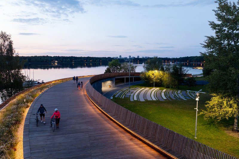 Cyclists and pedestrians on the timber boardwalk with amphitheater seating and illuminated tree at dusk