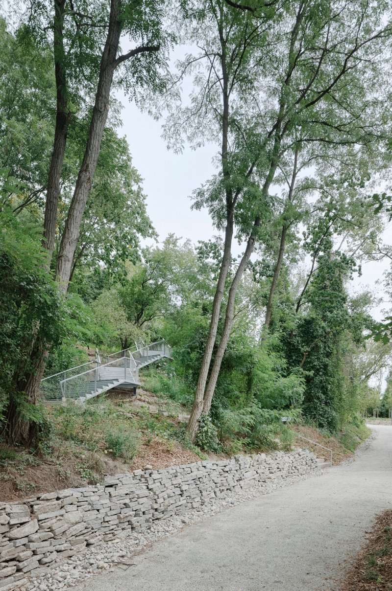 Metal footbridge ascending hillside amid trees beside a stone gabion wall and paved path