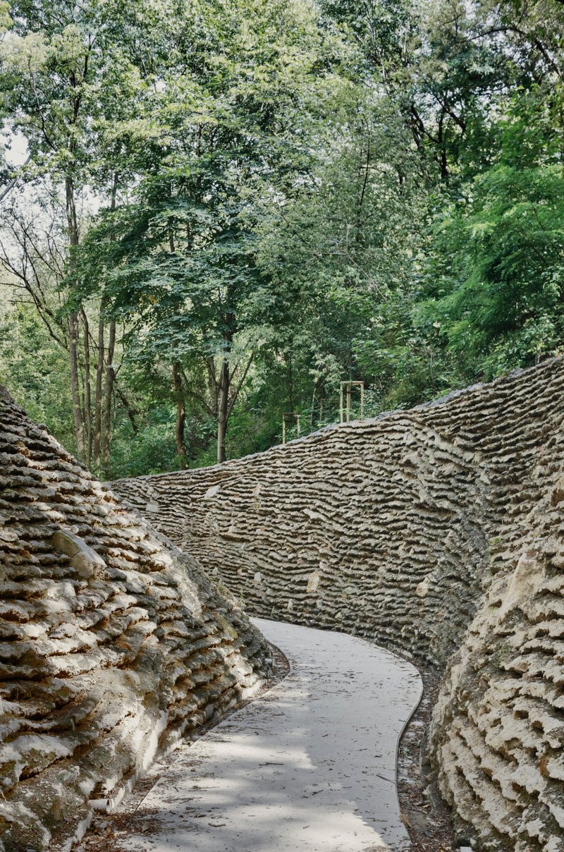Curving pathway enclosed by textured stone walls with rippled horizontal patterns beneath a canopy of trees