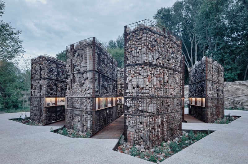 Cluster of cubic gabion volumes filled with rubble stone and integrated horizontal window openings at dusk
