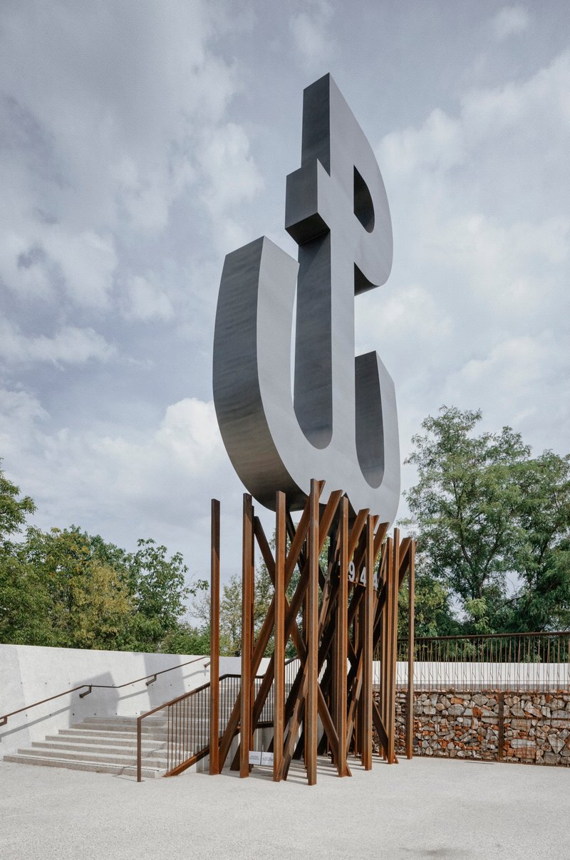 Sculptural metal letterform monument on vertical timber base beside concrete ramp and gabion retaining wall under cloudy sky