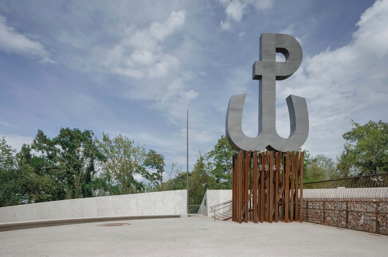 Metal letterform sculpture mounted on weathered timber slat screen beside concrete plaza and gabion wall with trees beyond