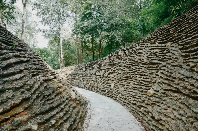 Curved concrete walkway enclosed by textured stone retaining walls with horizontal stratified layering under forest canopy