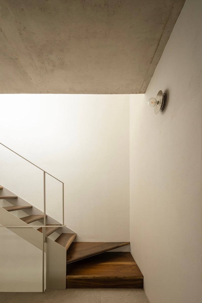 Wood staircase turning upward along white wall with circular sconce and concrete soffit above